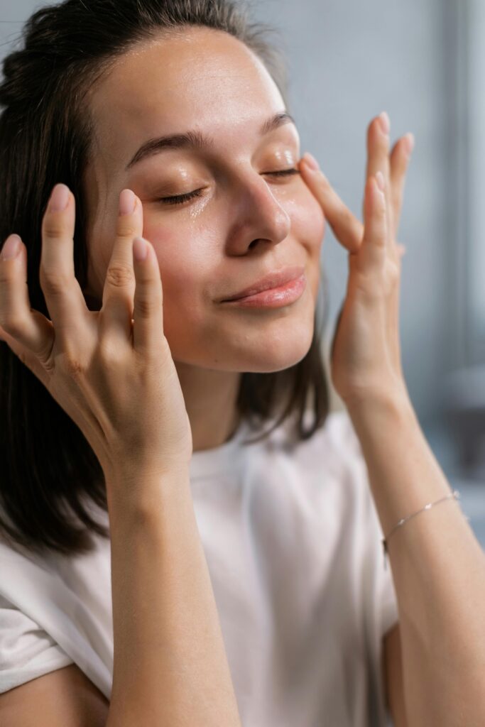 A woman with closed eyes gently massages her face, conveying relaxation and self-care. She wears a white shirt and her expression is serene.
